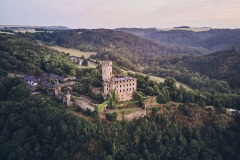Drohnenaufnahme einer mittelalterlichen Burg mit markantem Rundturm auf einem felsigen Höhenzug. Die Anlage ist von dichten Wäldern, Wiesen und sanften Hügeln umgeben und liegt eingebettet in einer ruhigen, ländlichen Landschaft bei weichem Abendlicht.