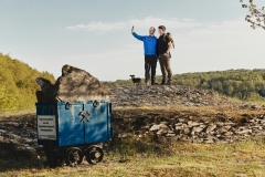 Zwei Wanderer stehen mit einem kleinen Hund auf einer Anhöhe und machen ein Selfie. Im Vordergrund befindet sich ein blauer Förderwagen aus dem Schieferbergbau mit einem großen Gesteinsblock. Die Landschaft ist von bewaldeten Hügeln und einem weiten Himmel geprägt.