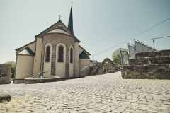 Die Pankratiuskirche in Kaisersesch präsentiert sich mit ihrer romanisch geprägten Apsis, hellen Natursteinfassaden und dem schlanken Kirchturm. Vor der Kirche erstreckt sich ein weitläufiger Platz aus geschwungen verlegtem Kopfsteinpflaster, ergänzt durch historische Mauerreste und eine Brunnenanlage.