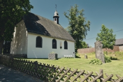 Die Waldkapelle in Kaisersesch steht in einer ruhigen, grünen Umgebung unter hohen Bäumen. Das schlichte, weiß verputzte Gotteshaus mit dunklem Satteldach und kleinem Glockenturm wird von einer gepflegten Rasenfläche, einem Holzzaun und einem Gedenkstein im Vordergrund eingerahmt.