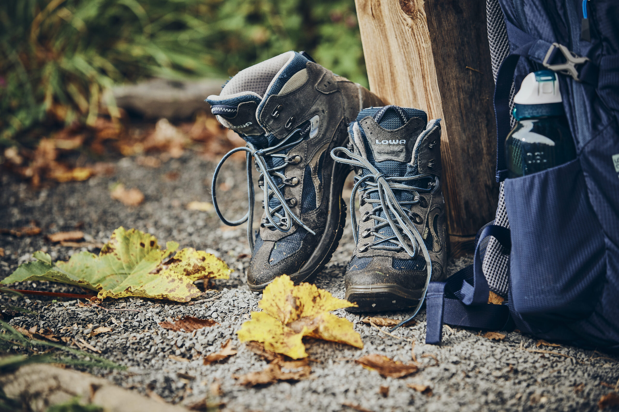 Wanderschuhe und ein Wanderrucksack sind an einen Holzstamm, auf einem Kieselweg, angelehnt. Ein paar Blätter liegen auf dem Weg und im Hintergrund sieht man Gras und Büsche.