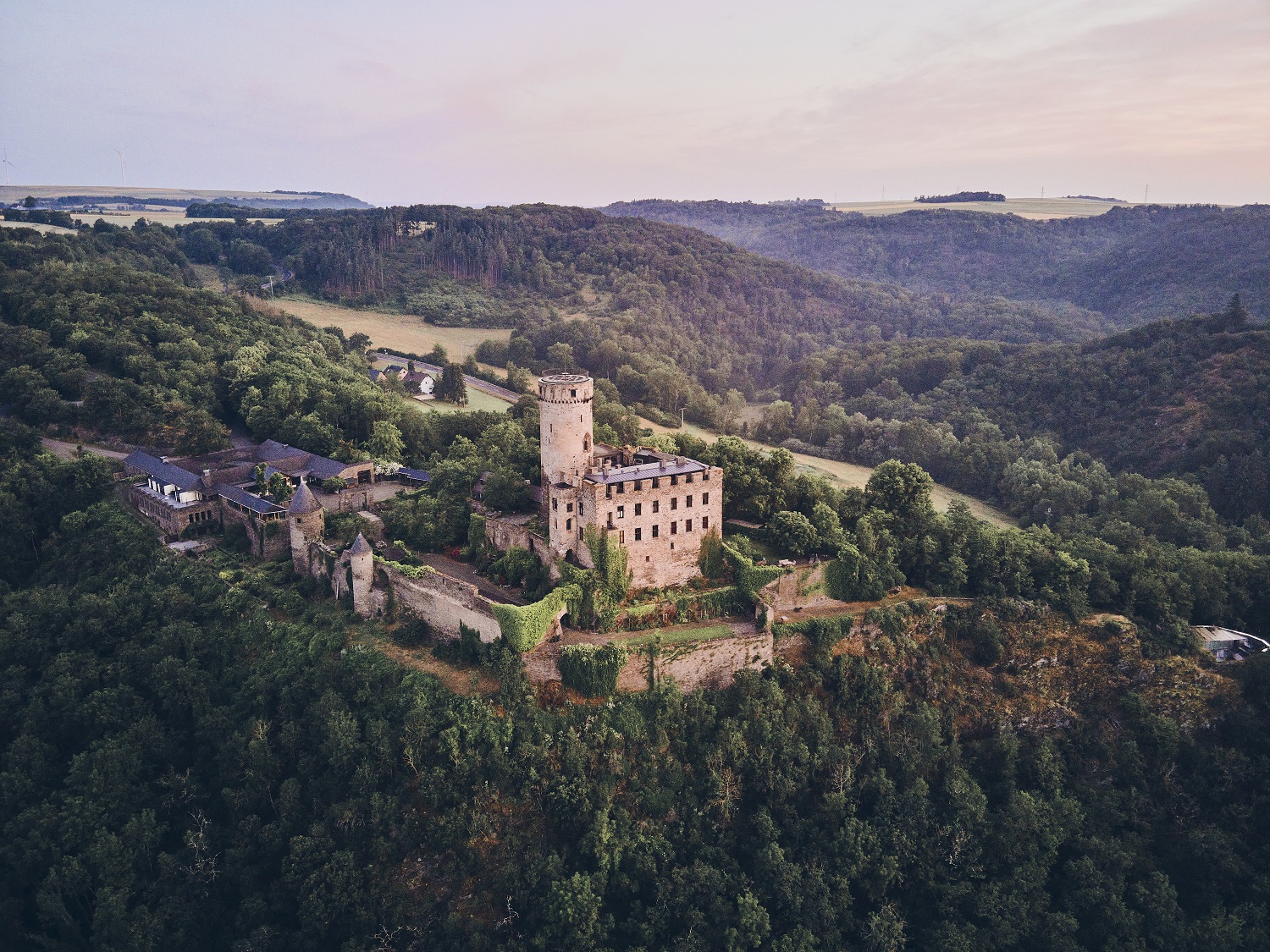 Drohnenaufnahme einer mittelalterlichen Burg mit markantem Rundturm auf einem felsigen Höhenzug. Die Anlage ist von dichten Wäldern, Wiesen und sanften Hügeln umgeben und liegt eingebettet in einer ruhigen, ländlichen Landschaft bei weichem Abendlicht.