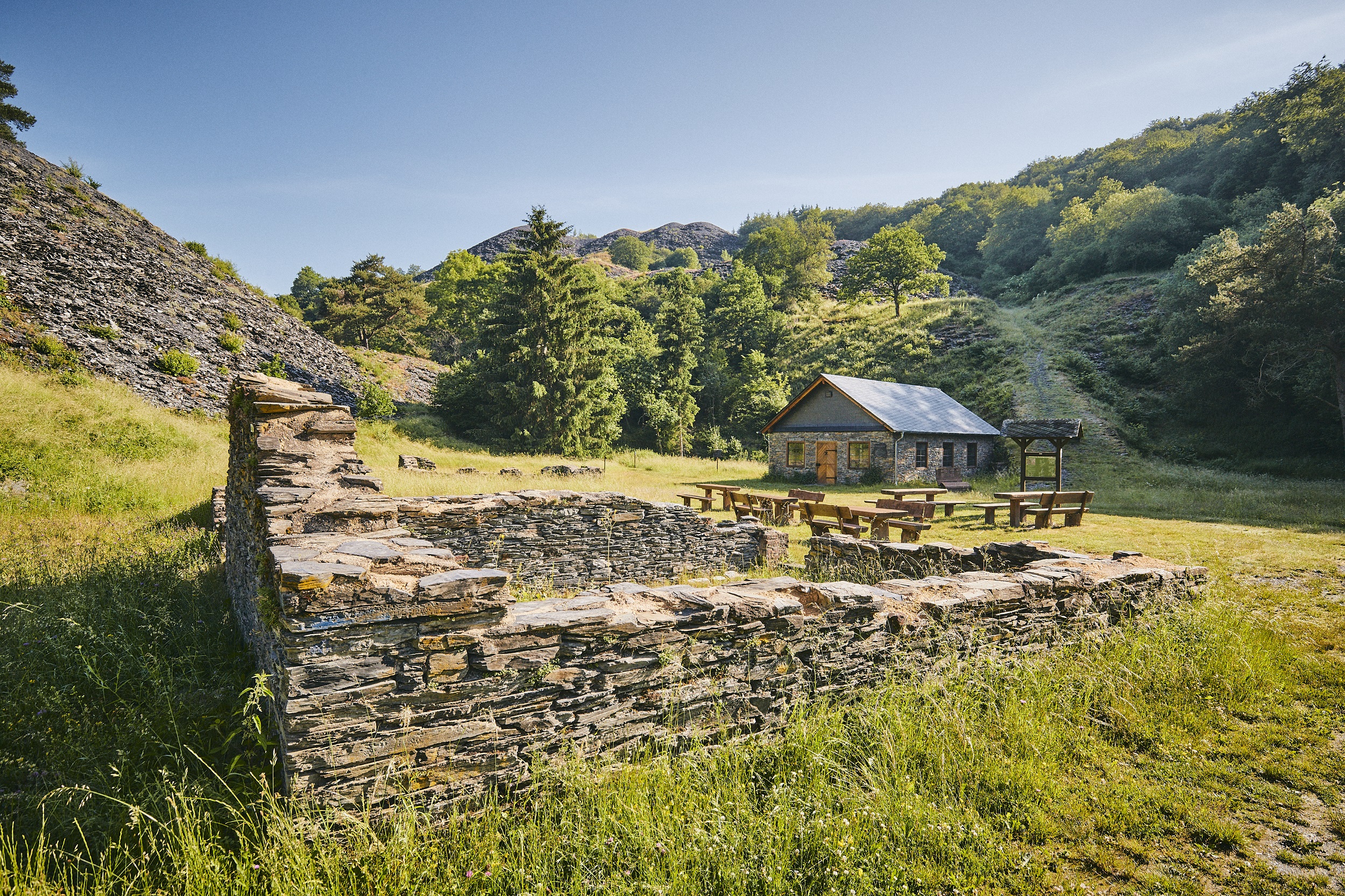 Ein Steinhaus mit Holzbänken auf einer Wiese, umgeben von grünen Hügeln und Bäumen