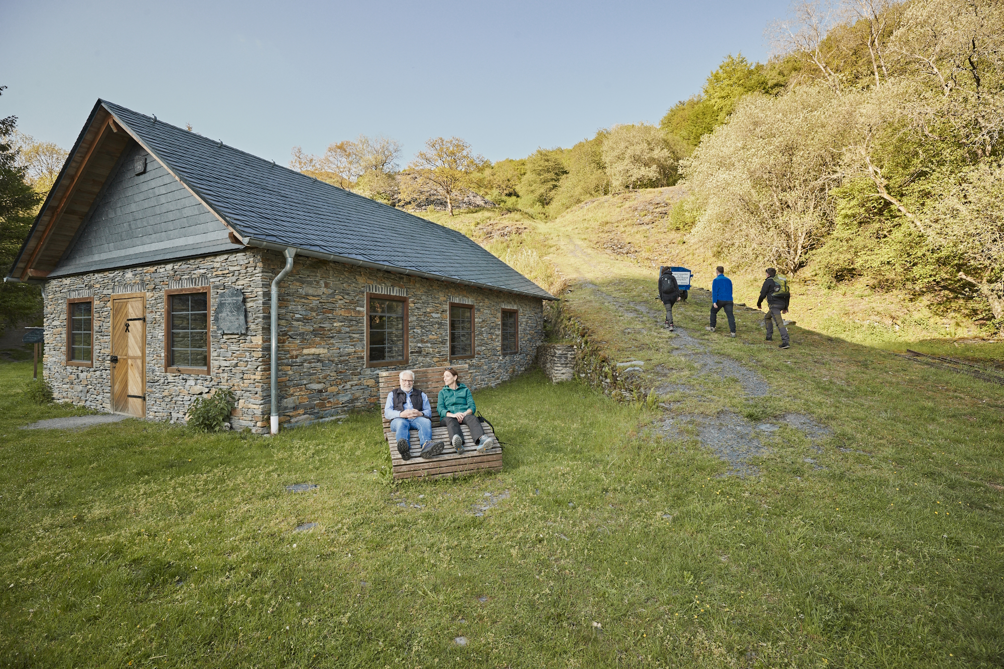 Zwei Personen sitzen auf einer hölzernen Liege auf einer Wiese vor einem historischen Schiefersteinhaus. Rechts führt ein leicht ansteigender Schotterweg den grünen Hang hinauf, den drei Wanderer bergauf gehen.