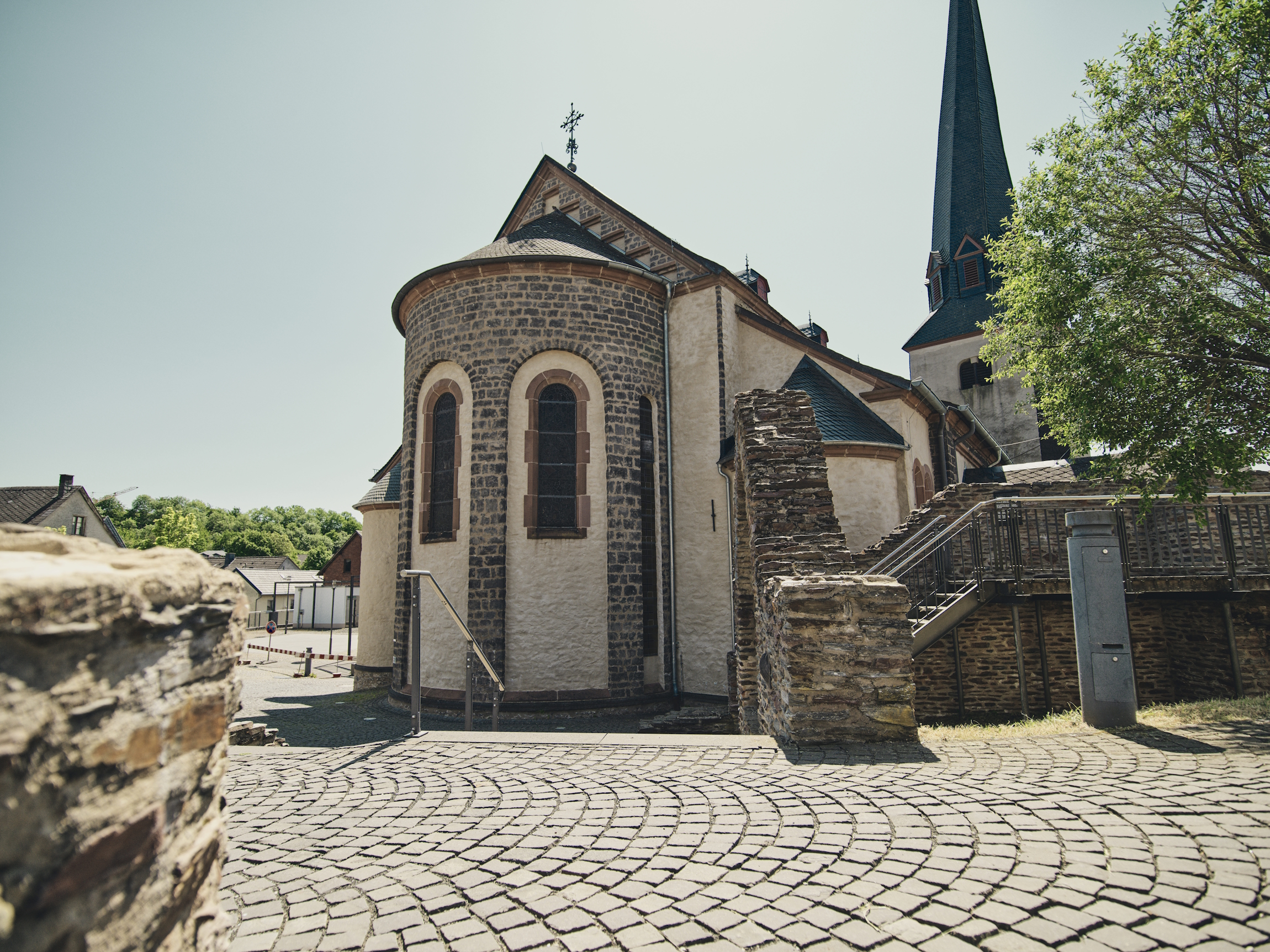 Historische Kirche mit markantem Turm, Natursteinmauerwerk und gotischen Fenstern an einem sonnigen Tag. Im Vordergrund verläuft ein gepflasterter Platz.