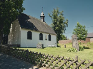 Weiße kleine Dorfkirche mit steilem Schieferdach und Turm, umgeben von Bäumen und einer gepflegten Rasenfläche. Im Vordergrund steht ein niedriger Holzzaun, rechts vor der Kirche ein steinernes Denkmal.